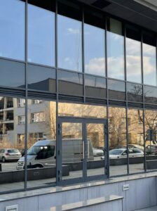 Przeszklona fasada biurowca z foliami lustrzanymi odbijającymi zaparkowane samochody i okoliczne budynki.
